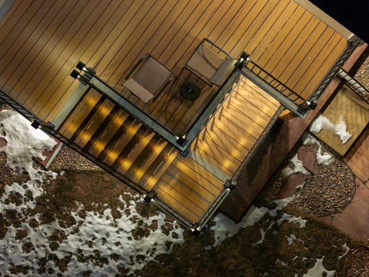 Nadir overhead view of a completed wood deck with illuminated LED stair risers, composite decking, metal railings, and snow surrounding the structure
