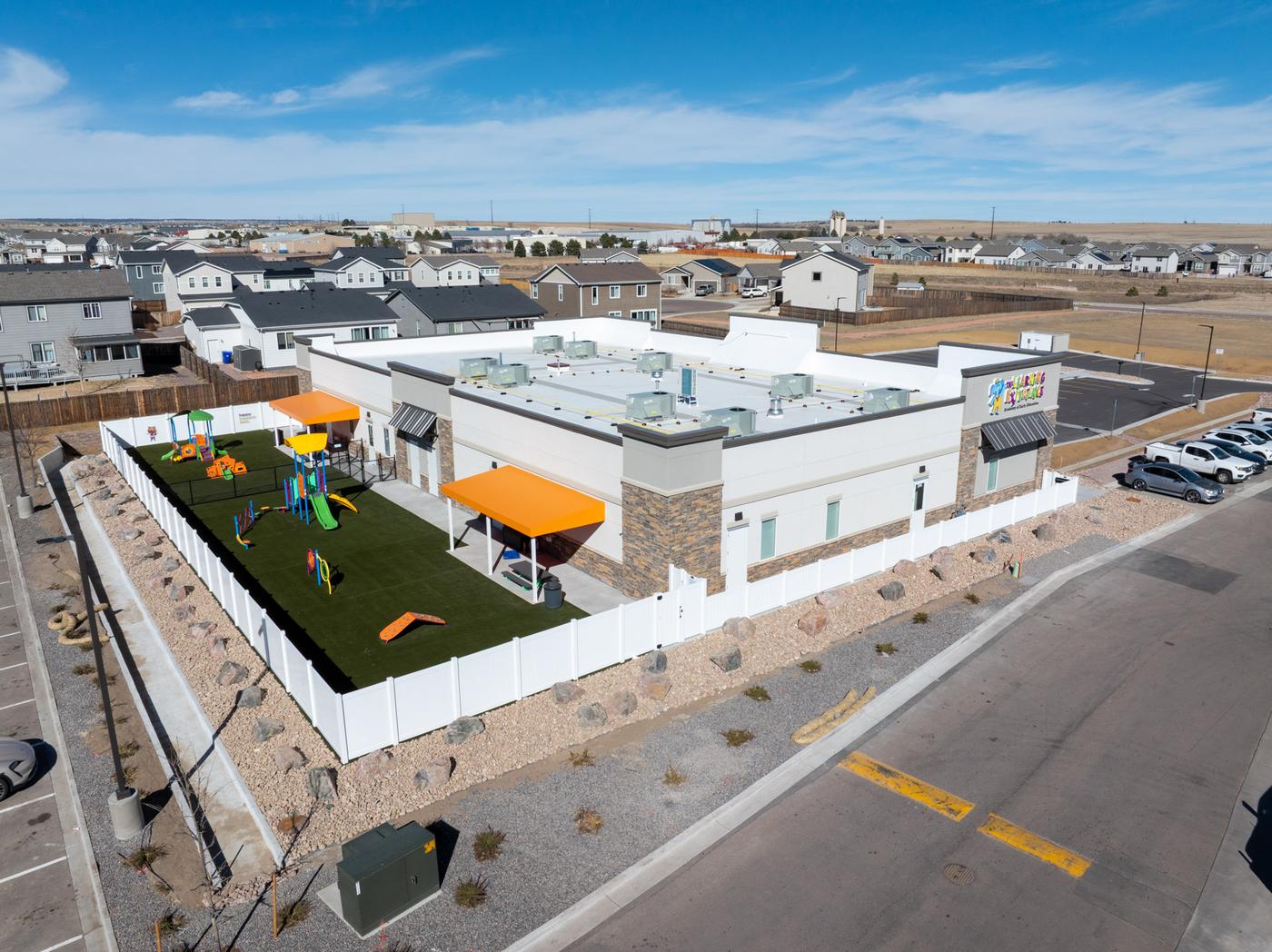 Aerial view of a newly completed commercial childcare facility with colorful playground equipment and orange shade canopies in the fenced yard, modern flat-roof construction, Colorado Springs