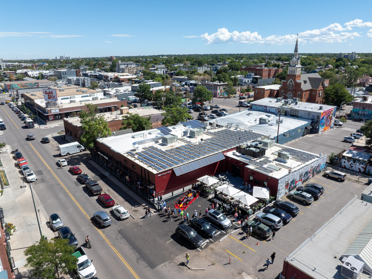 Medium altitude aerial view looking down on the F1 Arcade venue in Denver's RiNo neighborhood, with a Red Bull Formula 1 car visible in the parking lot surrounded by event attendees