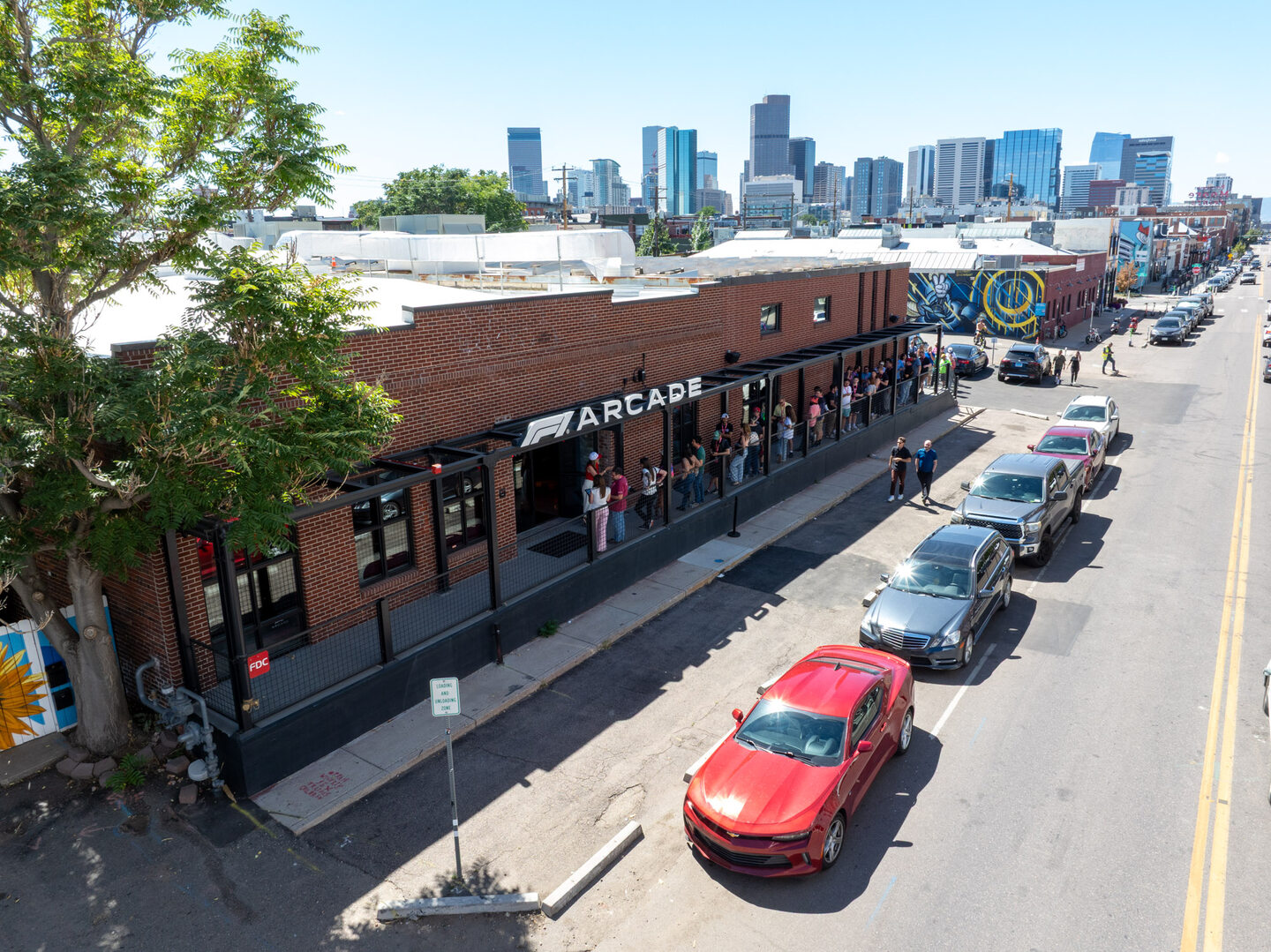 Low aerial view of the F1 Arcade building facade with a queue of attendees on the terrace, Denver city skyline and the Rocky Mountain Front Range visible behind