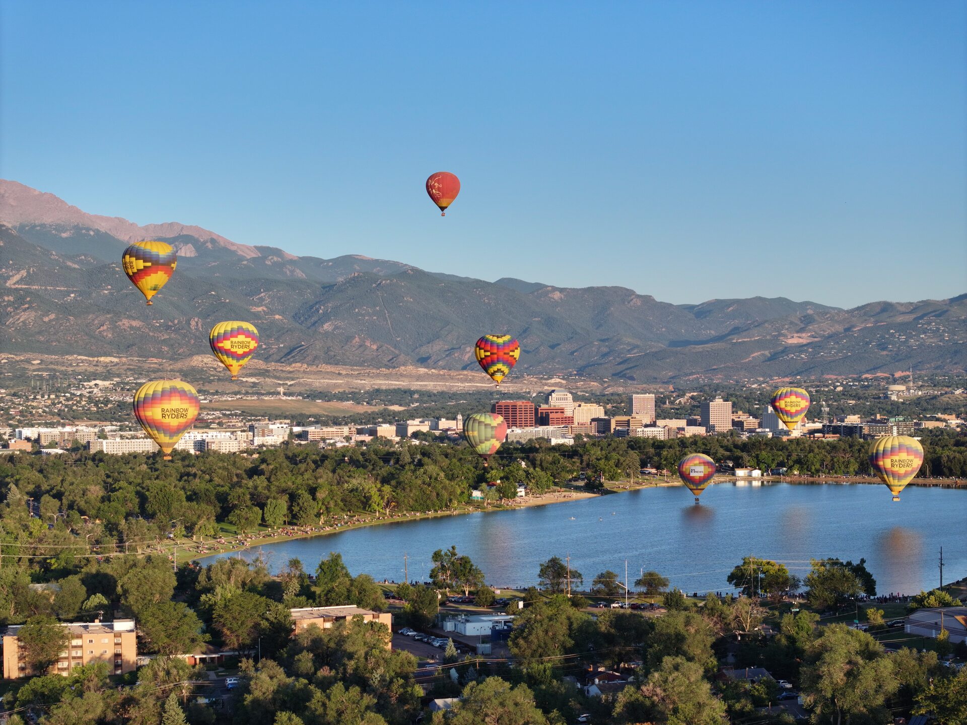 Golden sunrise aerial view of hot air balloons launching at dawn during the 2024 Labor Day balloon classic at Prospect Lake Park, Colorado Springs, with warm morning light across the city