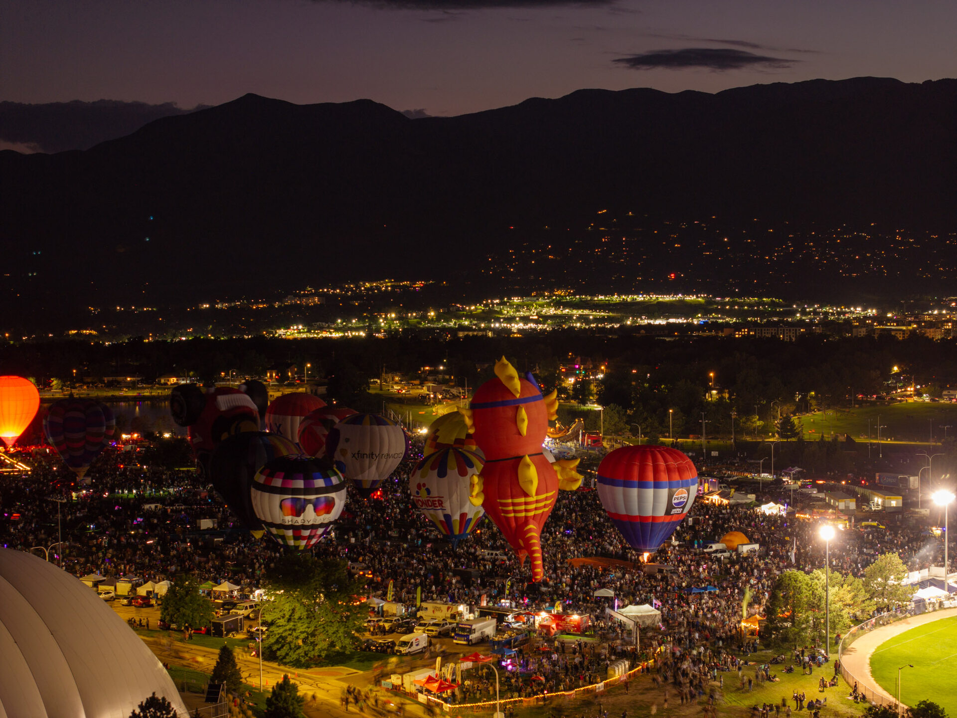 Wide night aerial view of the Labor Day balloon glow festival over Colorado Springs, with glowing hot air balloons, tens of thousands of attendees, Cheyenne Mountain silhouetted, and city lights stretching to the horizon