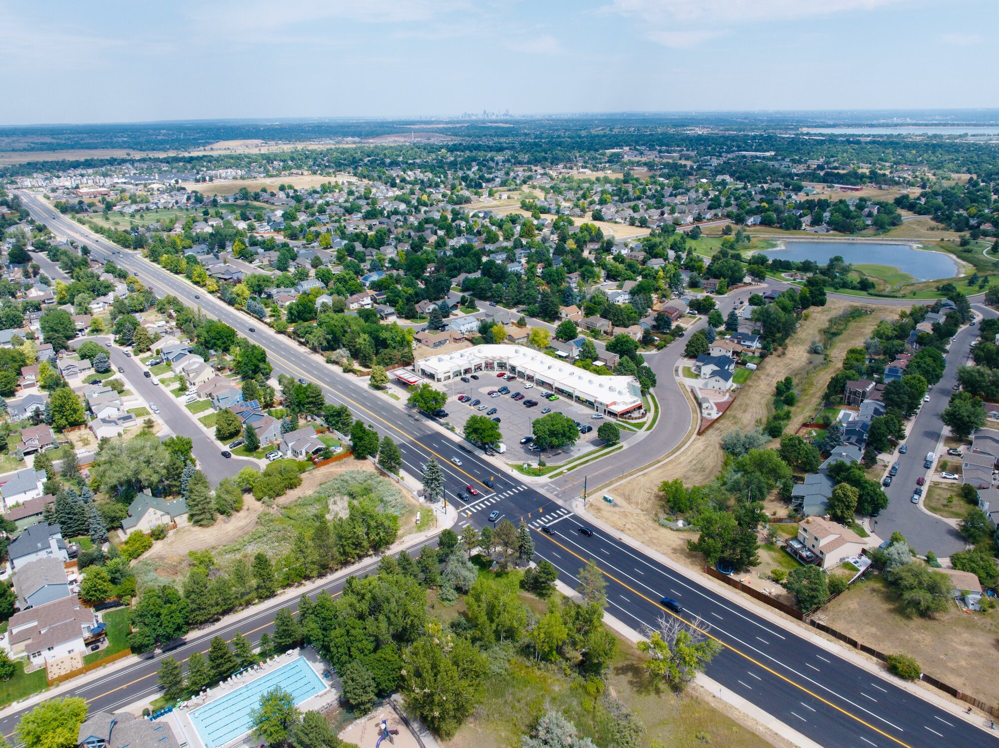 Aerial view of an established Colorado residential suburb — community pool, lake, mature trees, and residential streets visible with the city skyline on the horizon
